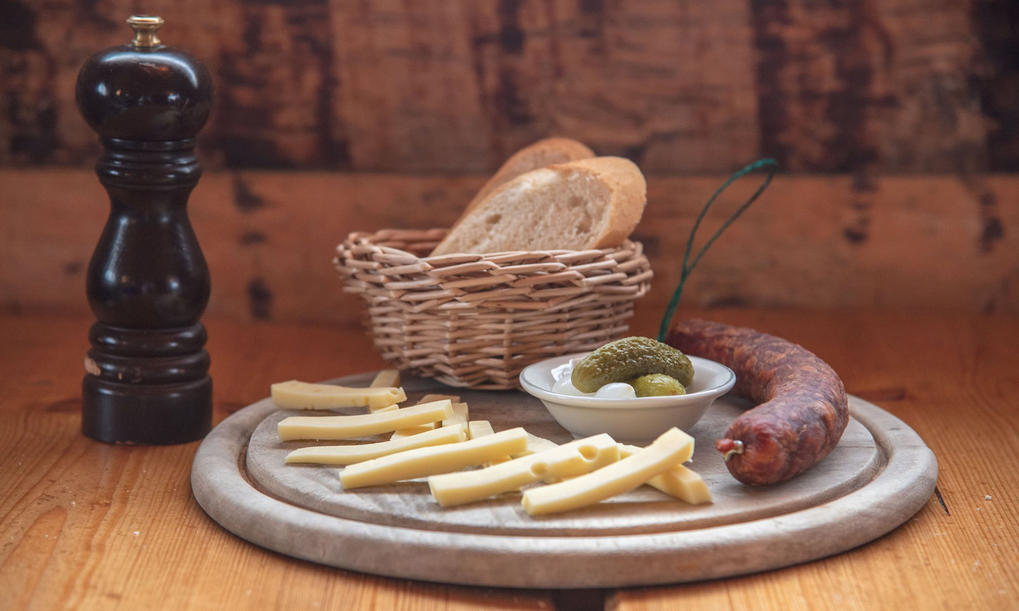 Photo d'une assiette apéritive, sur les bord du fromage et un saucisson entier, au centre un petit bol de cornichon et oignon et au fond un panier de pain