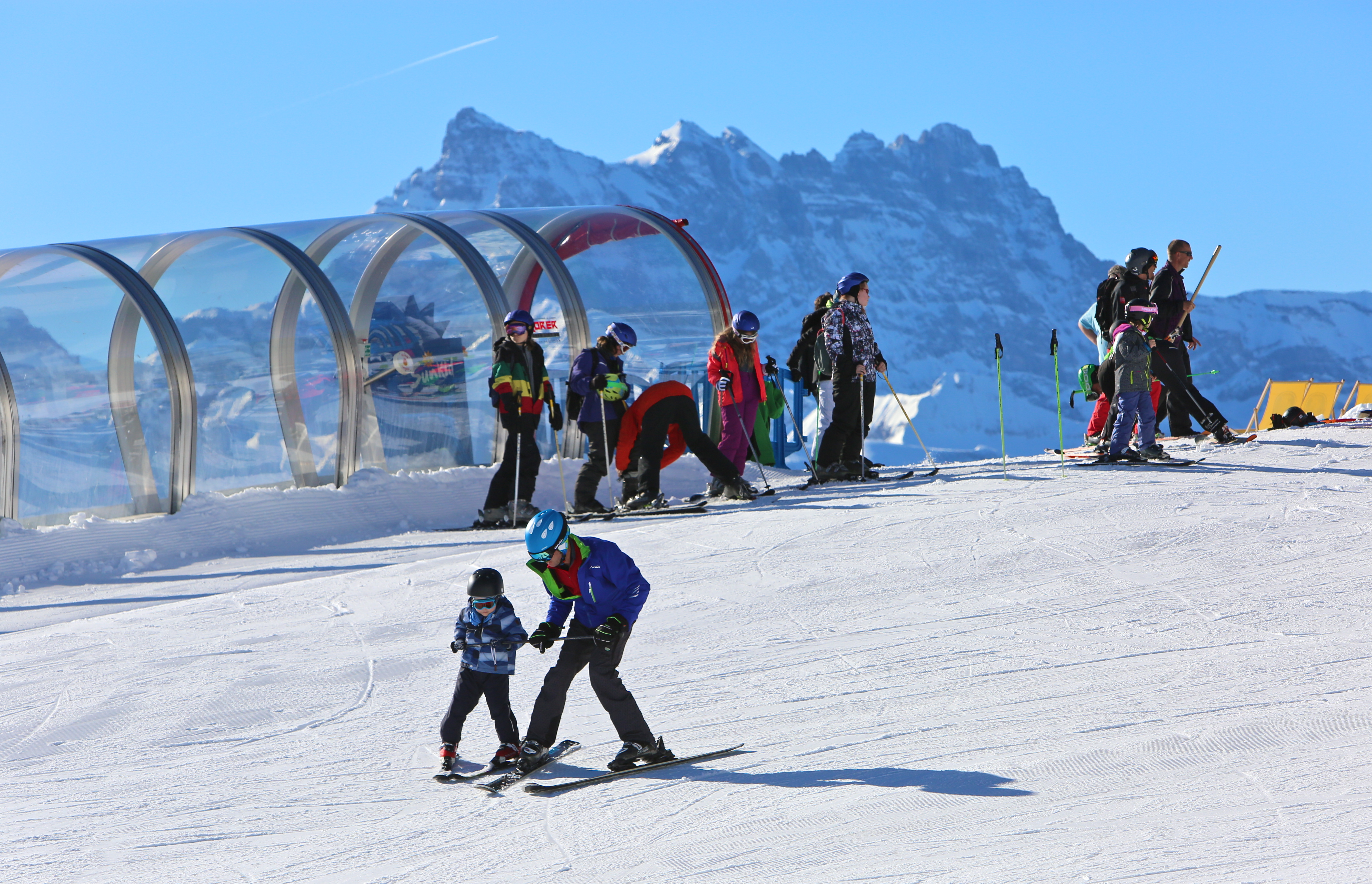 Enfant apprenant à skier à la vallée blanche avec un adulte. En arrière un tapis roulant couvert avec un groupe de personne à la fin. Dents du Midi en arrière plan.